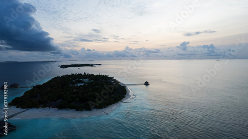 Obraz na płótnie Top view of the sunset over an island in the ocean. Large dark clouds with a orange tinge. Turquoise ocean water, white sand of the beach. Green palm trees. Villas on the water. Water is like a mirror w salonie