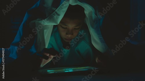 A boy is sitting in a dark room with a tablet in his hand. He is looking at the screen with a serious expression