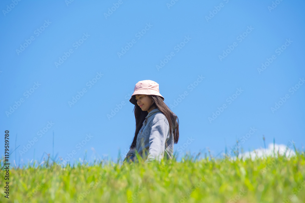 Young Asian woman walking on a green hill, smiling at the camera, against a blue sky Young Asian woman walking on a green hill, smiling at the camera, against a blue sky