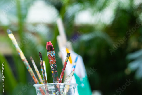 Selective focus with copy space of colorful paint brushes in a cup, splattered with paint, against a lush, blurred backdrop for creativity, art supplies, and artistic expression in nature outdoor.