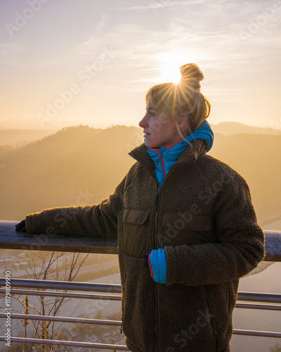 Tourist girl in warm clothes stands on Bastay Bridge in Saxon Switzerland at sunset, enjoying view of majestic cliffs, Elbe River. Popular tourist destination. Germany. photo