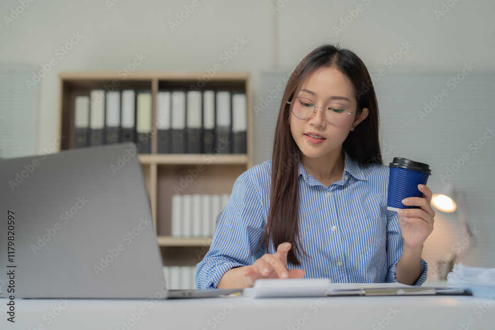 Fototapeta premium Asian businesswoman wearing glasses, working intently on a laptop while sipping coffee at her office desk, balancing professional tasks in a modern corporate environment