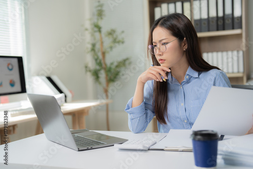 Asian businesswoman concentrating on analyzing financial documents and working with laptop computer at office desk, thoughtful accountant reviewing financial reports photo