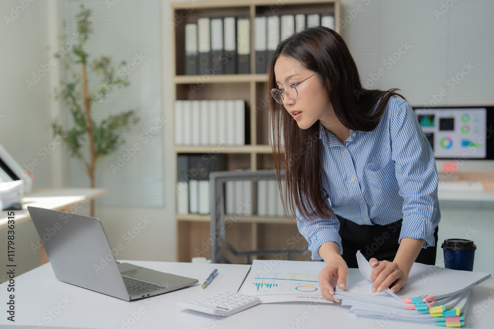 Businesswoman wearing glasses is searching through a stack of documents at her desk, with a laptop, calculator, and charts also present, indicating a busy and productive work environment Businesswoman wearing glasses is searching through a stack of documents at her desk, with a laptop, calculator, and charts also present, indicating a busy and productive work environment