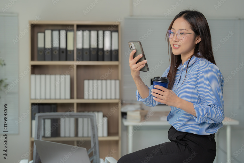 Young Asian businesswoman wearing glasses using smartphone and drinking coffee while sitting on desk in modern office, shelves with folders in background Young Asian businesswoman wearing glasses using smartphone and drinking coffee while sitting on desk in modern office, shelves with folders in background