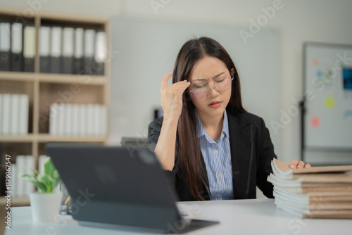 Young businesswoman experiencing a headache while working on a laptop at her office desk, reflecting the challenges of chronic work stress and fatigue