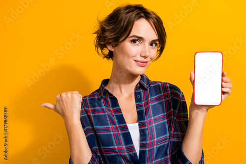 Photo of charming woman dressed plaid shirt show smartphone display indicating at eshop empty space isolated on yellow color background photo
