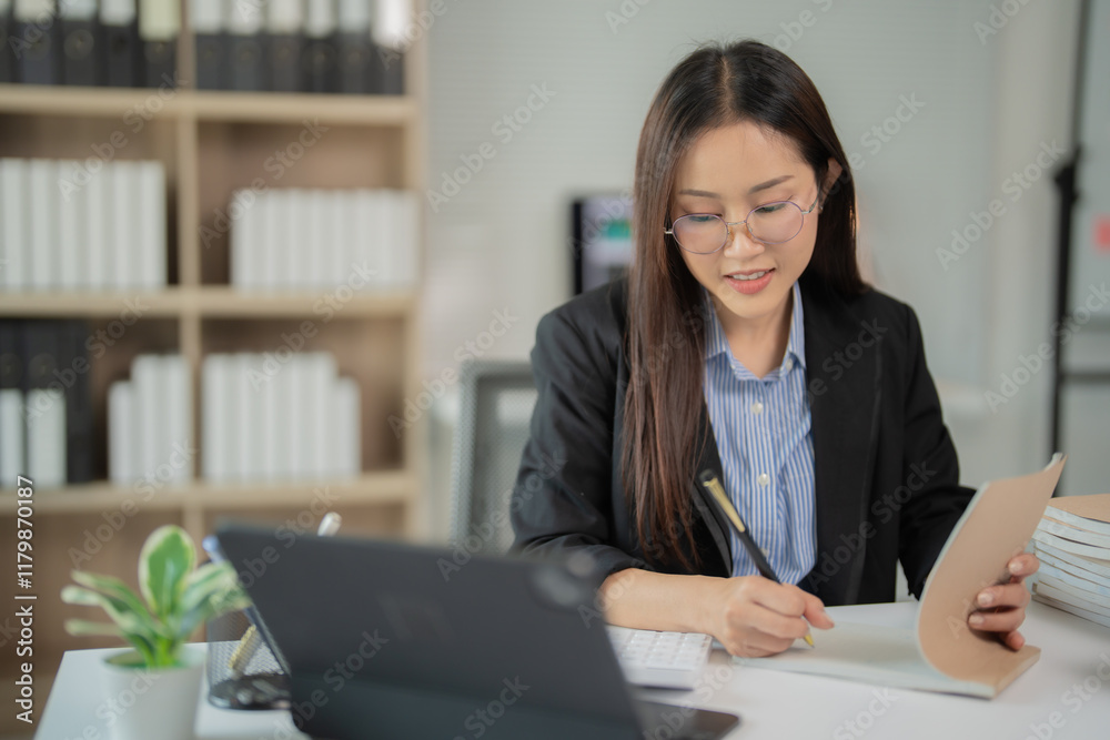 Asian businesswoman wearing glasses and suit sitting at desk taking notes in notebook while using tablet and keyboard in office with bookshelves in background Asian businesswoman wearing glasses and suit sitting at desk taking notes in notebook while using tablet and keyboard in office with bookshelves in background
