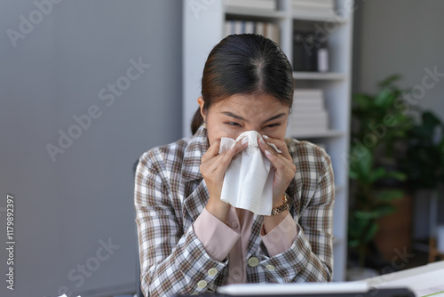 Asian businesswoman blowing her nose into a tissue while working at her desk in the office, dealing with symptoms of a cold or allergy
