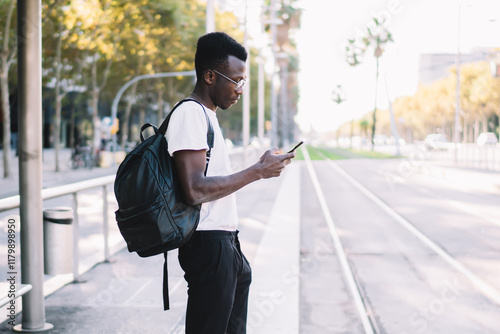 Handsome trendy dressed male tourist checking navigation via application on smartphone with trafic cand public transport schedule. young african american man waiting for bus on station use cellular photo