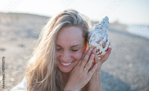 Smiling pretty blonde listening the seashell at the beach #1179938776