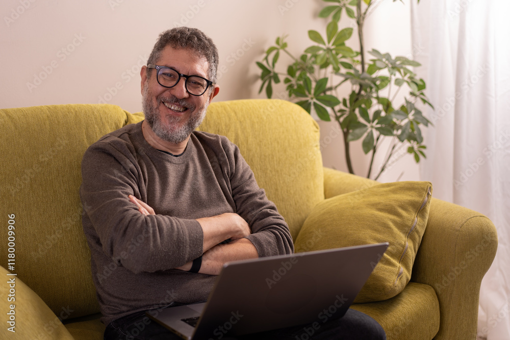 Mature man smiling while seated on yellow sofa with laptop Mature man smiling while seated on yellow sofa with laptop