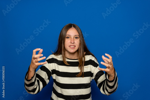 Beautiful hispanic teenage girl about 15 years old wearing a striped sweater with clapped hands in a sign of rage and anger, isolated on blue studio background.