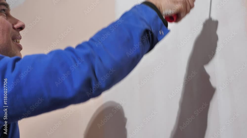 Side view of cheerful builder painter painting wall using roller and white acrylic paint. Smiling builder during work indoors. Side view of cheerful builder painter painting wall using roller and white acrylic paint. Smiling builder during work indoors.
