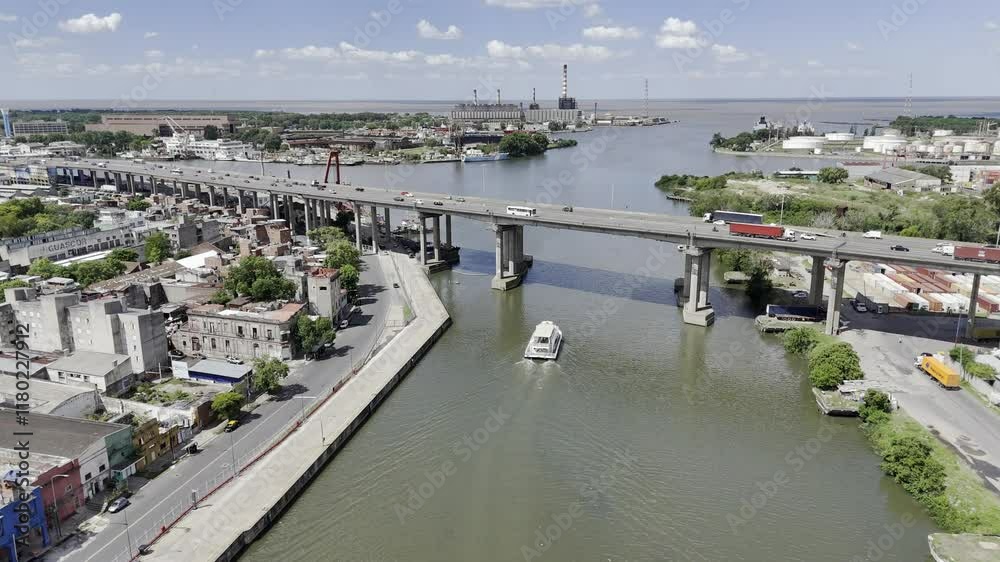 Drone flies over red bridge toward concrete bridge crossing river, with lighthouse and the sea in the distance