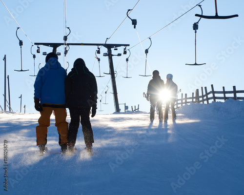 Persons riding a t-bar lift upwards a skiing slope in a winter landscape backlit by the sun shining though blowing snow