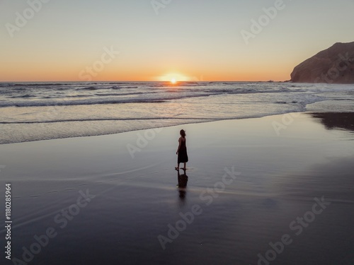 Fototapeta Naklejka Na Ścianę i Meble -  Solitude at sunset. Woman in dark dress walks along tranquil beach at golden hour. Peaceful reflection. Bethells Beach, Auckland, New Zealand