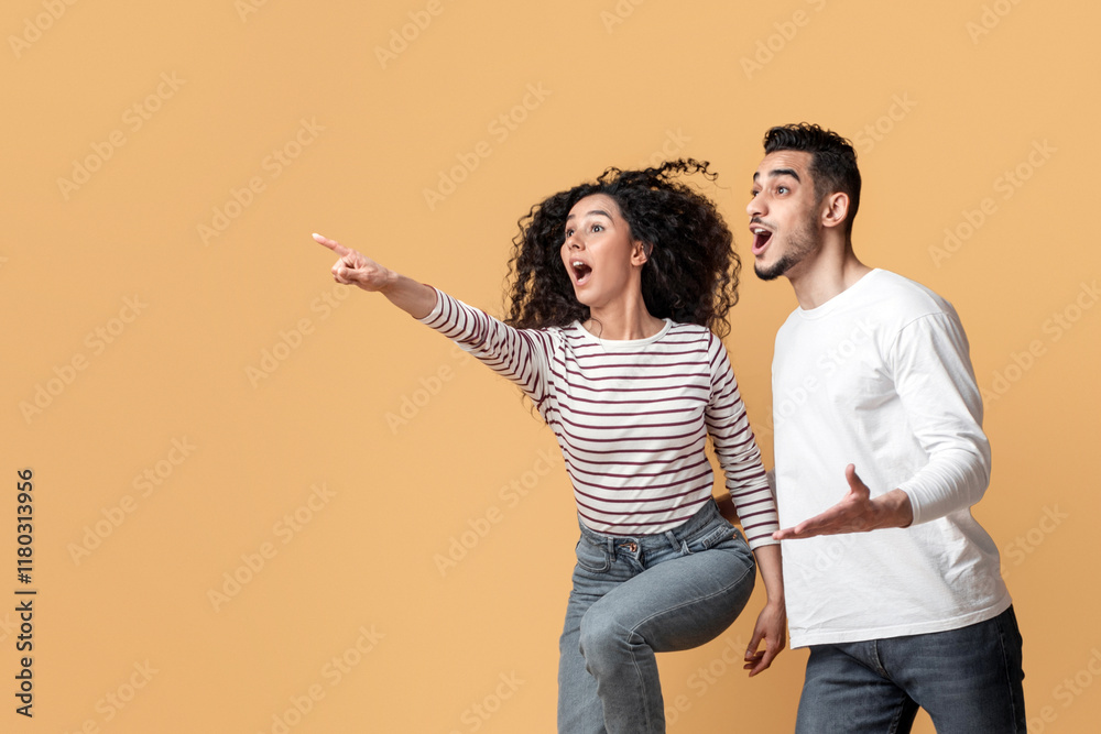 Look There. Amazed Arab Couple Pointing Aside While Jumping On Yellow Background, Excited Young Man And Woman Demonstrating Copy Space For Design Or Advertisement, Full Length Look There. Amazed Arab Couple Pointing Aside While Jumping On Yellow Background, Excited Young Man And Woman Demonstrating Copy Space For Design Or Advertisement, Full Length