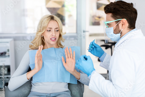 Scared blonde woman female patient sufffering from dental phobia, sitting at dental chair, looking at tools in dentist hands and raising hands in stop or no gesture, avoid treatment