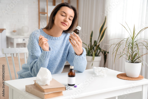 Sick young woman with cough syrup at table in bedroom