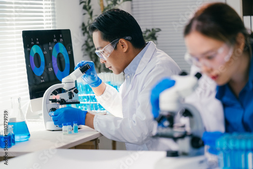 Two medical researchers wearing lab coats and safety glasses are using microscopes and analyzing data on computer screen while developing new treatments in modern laboratory