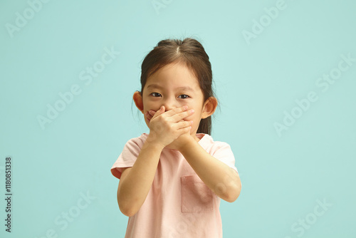 Ooops, i said it. Naughty korean Child Girl with Hand Over Mouth, does not want to tell the truth, tries to deceive or keep silent about something standing over blue isolated background.