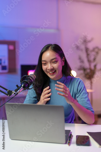 Asian woman smiles while recording a podcast in her home studio using a professional microphone and laptop, gesturing with her hands as she engages with her audience in a live stream
