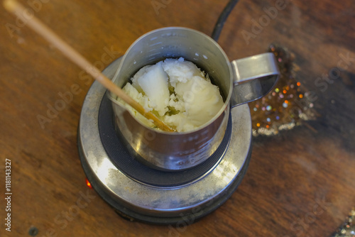 Close-up of an iron bowl on a stove where beeswax is melted to make candles. The woman stirs the wax periodically. The process of making a handmade scented candle in a home workshop. photo