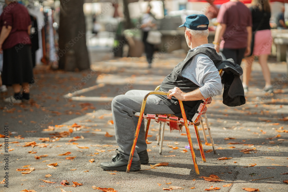 Older unknown person sitting in a chair and enjoying the hustle of an open street market. Elder person relaxing in an old colorful beaten up chair.