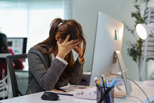 Young professional woman experiencing stress and burnout while working on a computer in a modern office, covering her face with hands and struggling with a difficult task or deadline