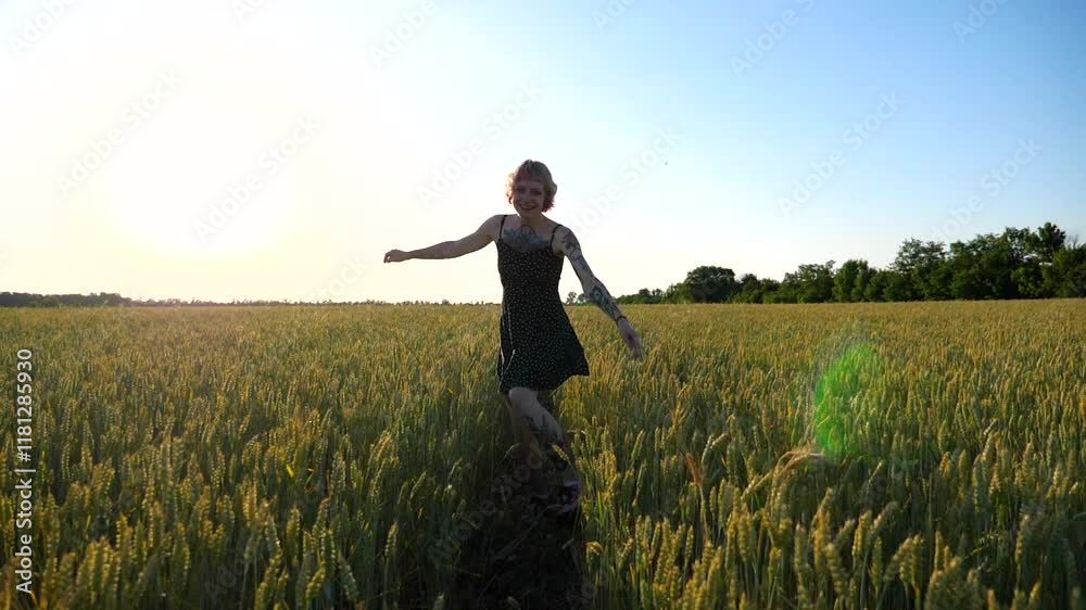 Cheerful hippie girl with pink hair having fun while running among green barley field. Happy punk woman with tattoos jogging through wheat meadow at sunset. Joyful female hipster enjoying summer