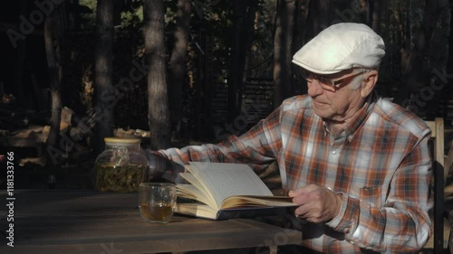 An elderly man in a flat cap pours tea into a cup while seated at a wooden table, immersed in a book amidst a serene forest backdrop on a sunny afternoon.