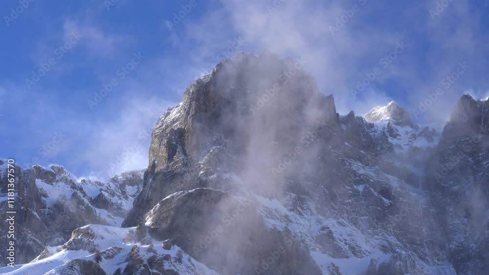 Strong wind blows the snow around on the mountain peaks in winter. The snow swirls up and winds over the edge. It is an impressive, wild landscape. It's cold and the sky is blue. sun shines on snow Strong wind blows the snow around on the mountain peaks in winter. The snow swirls up and winds over the edge. It is an impressive, wild landscape. It's cold and the sky is blue. sun shines on snow