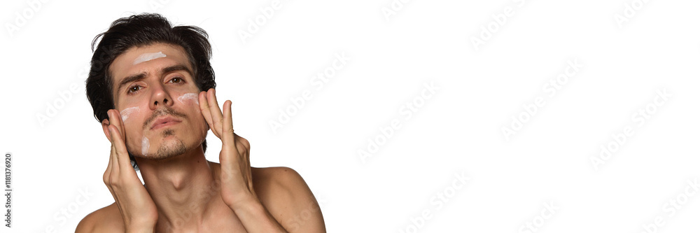 Banner. Man applying cream on face, concentrated on facial care routine against white background with negative space to insert text. Concept of male beauty and facial care, cosmetology. Banner. Man applying cream on face, concentrated on facial care routine against white background with negative space to insert text. Concept of male beauty and facial care, cosmetology.