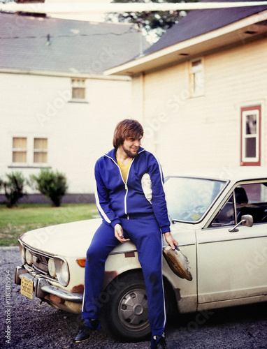 Young man in blue track suit with muddy shoes leaning against car in driveway
 photo