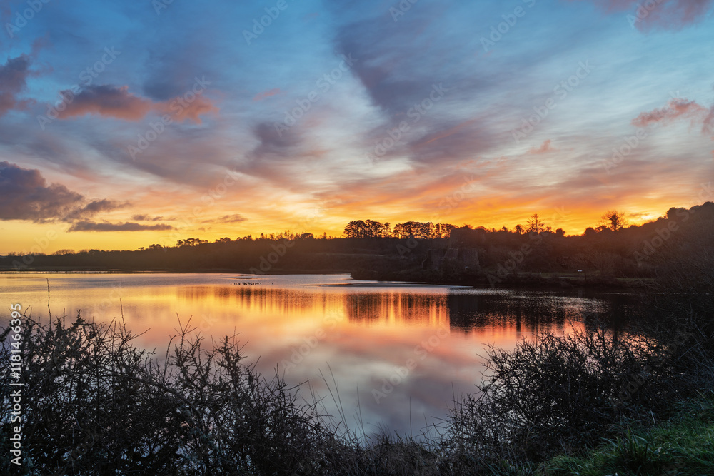 Fototapeta premium Lever de soleil aux teintes orangées se reflétant sur l'étang de l'Aber, presqu'île de Crozon. Quelques nuages se dessinent dans le ciel et se mirent dans l'eau paisible.