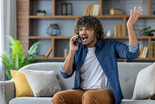 Angry and spiteful man talking on the phone while sitting on the couch in the living room at home, dissatisfied customer complaining to customer support service.