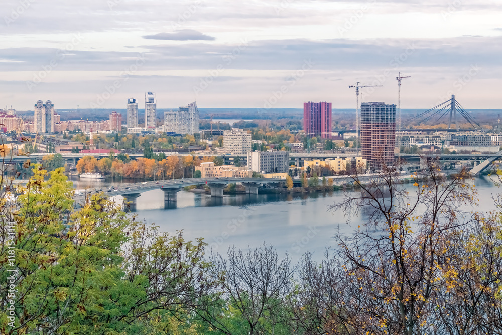 Fototapeta premium A tranquil view of Kyiv, Ukraine, showcasing the Dnipro River and urban skyline. Autumn foliage and city bridges add depth to the serene landscape