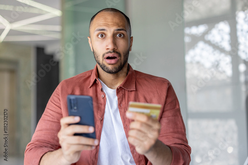 Portrait of disappointed and deceived man, holding bank credit card and phone in hands, account blocked and money transfer refused. Fraudulent actions.