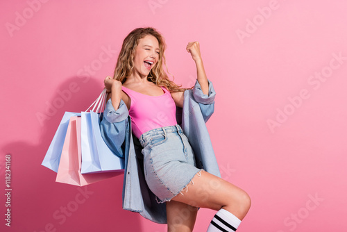 Joyful young woman in stylish denim outfit celebrating with shopping bags against pink background photo