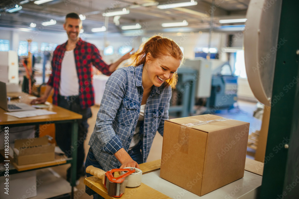 Female worker packing boxes in factory with colleague smiling in background Female worker packing boxes in factory with colleague smiling in background