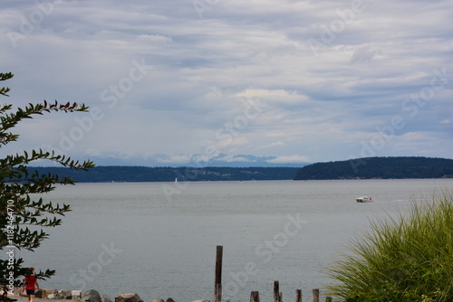 Serene waterfront view with a distant boat, lush greenery, and dramatic clouds over a tranquil bay surrounded by forested hills. photo