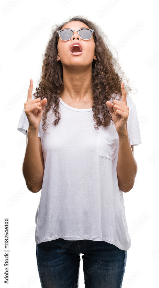 Naklejka premium Young hispanic woman wearing sunglasses amazed and surprised looking up and pointing with fingers and raised arms.