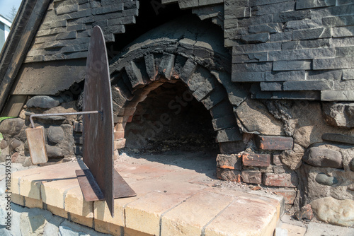 A vintage outdoor wood-fired bread oven. The exterior structure is made from brick and rocks with a circular heavy metal door and an iron and wood handle. There's black soot on the hearth of the stove photo