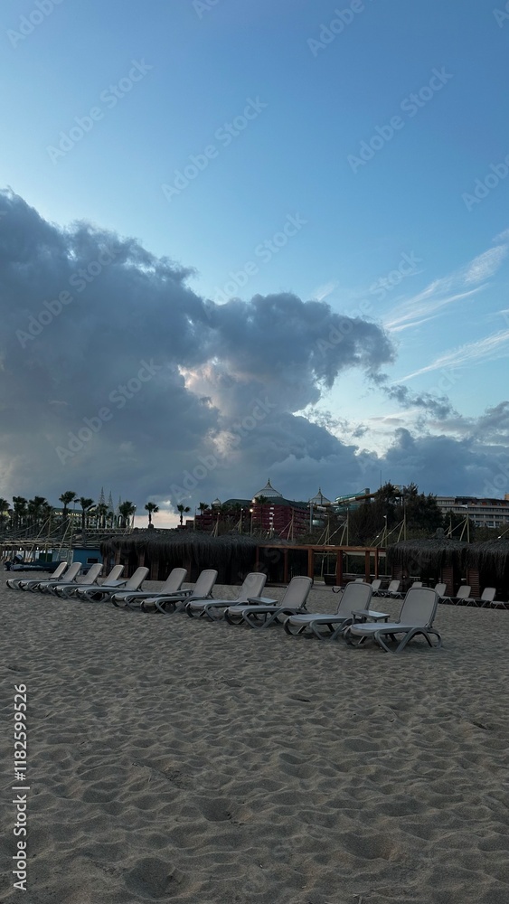 Obraz premium Empty sun loungers on a sandy beach with a dramatic sky, resort buildings, and palm trees in the background. Peaceful vacation scene with storm clouds and a tranquil seaside atmosphere