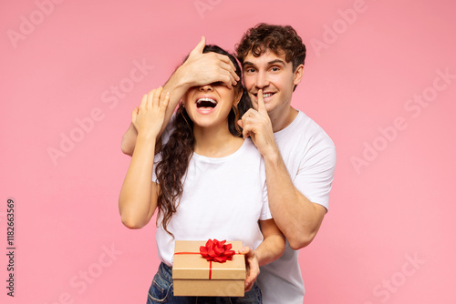 Happy guy closing eyes to wife, giving gift box and making shh silence sign over pink studio background. Surprise, anniversary, birthday, Valentine's day photo