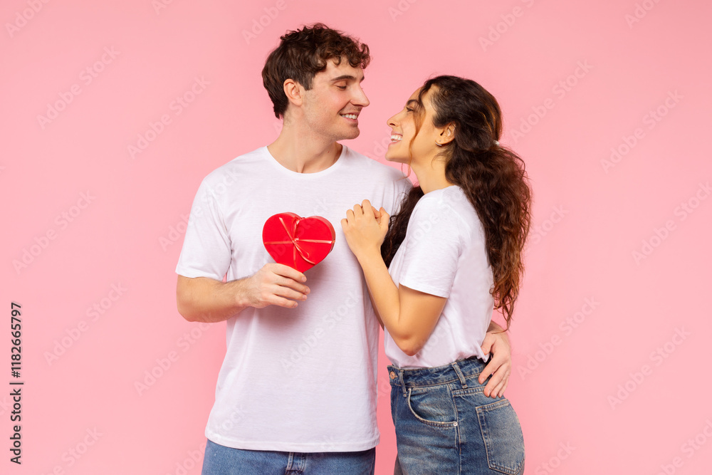 St. Valentines Day. Happy European couple in love embracing, man holding red heart shaped gift box, looking at each other, posing on pink studio background St. Valentines Day. Happy European couple in love embracing, man holding red heart shaped gift box, looking at each other, posing on pink studio background