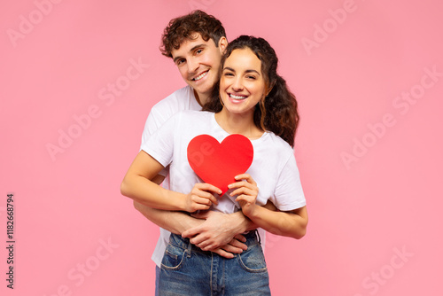 Young European couple in love embracing, holding paper heart, posing over pink studio background. Valentine's day celebration and romantic relationship