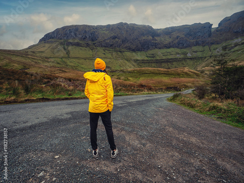 Travel and tourism in Ireland. Teenager girl standing on a side of a road and looking at majestic nature scenery of Gleniff Horseshoe drive with tall mountains. Model wearing yellow jacket and hat. photo