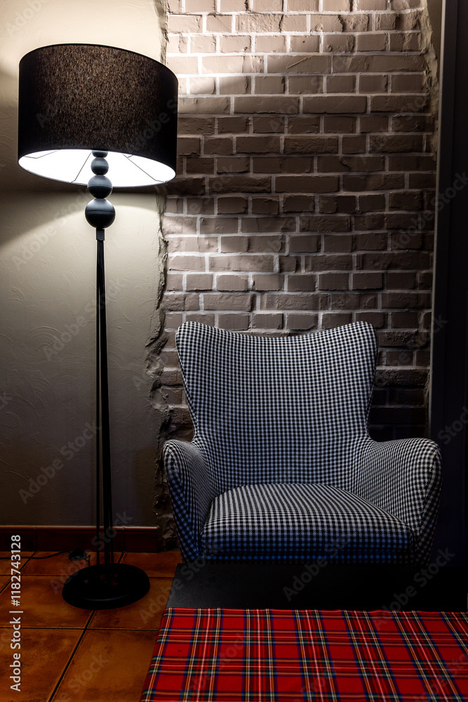 A grey, comfortable armchair with a houndstooth pattern stands against a grey brick wall. The scene is lit by the atmospheric light of a floor lamp.